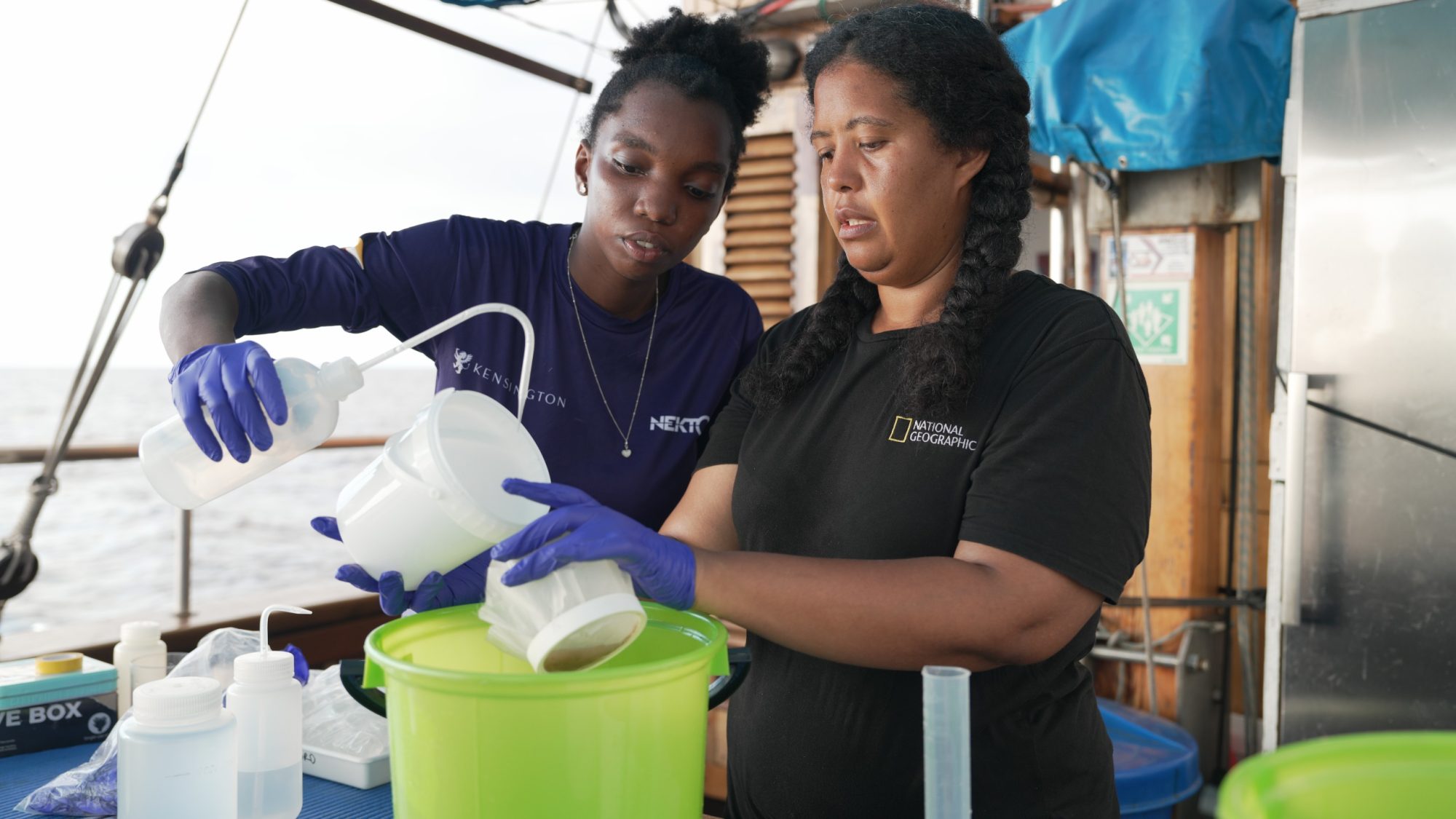 Two women work together on a boat, pouring liquid from a bottle into a container. They wear gloves and focused expressions, symbolizing collaboration in a scientific setting.