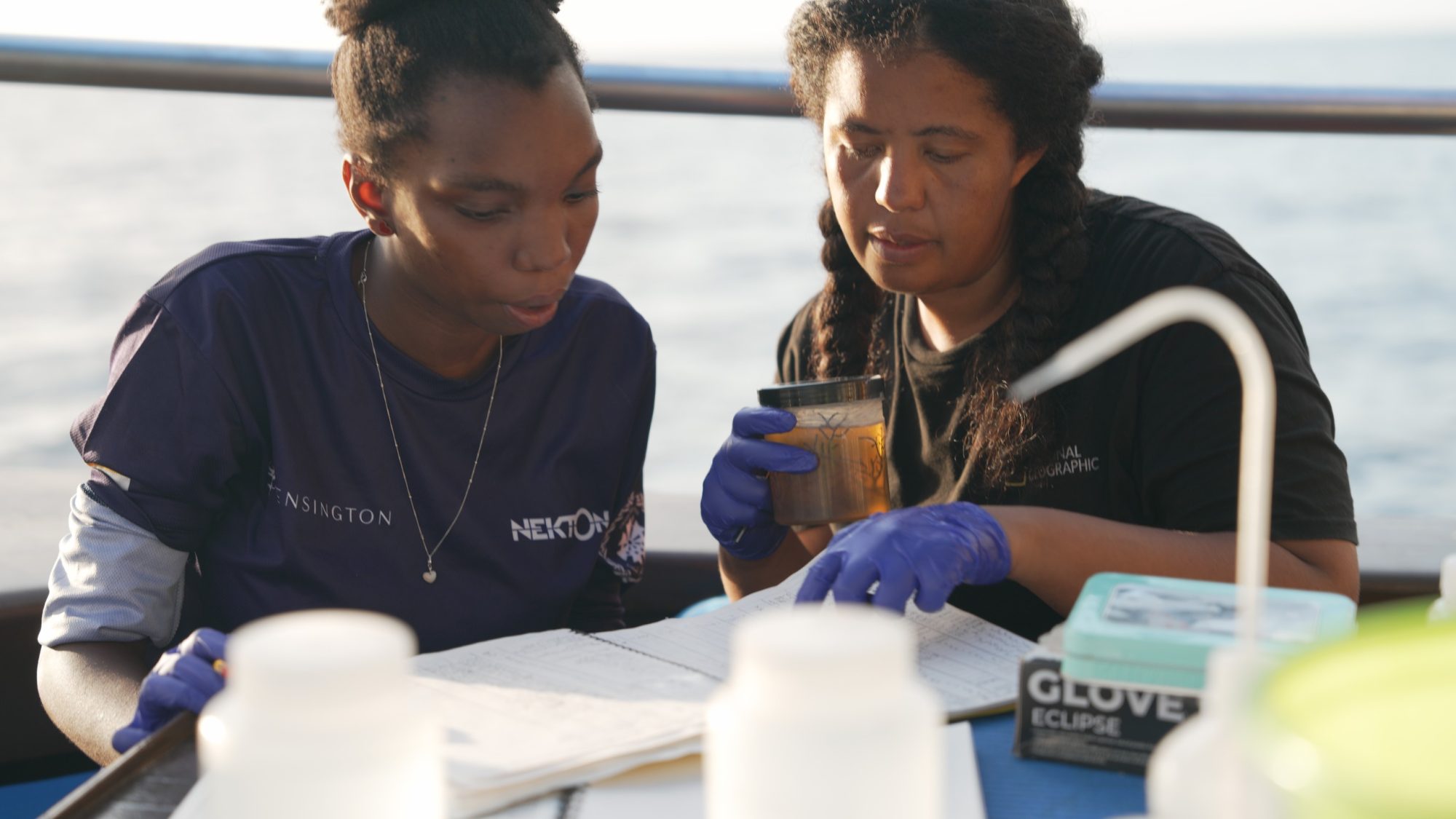 Two women on a boat examine a document. They wear blue gloves, conveying focus and collaboration. Laboratory items and the ocean visible in background.
