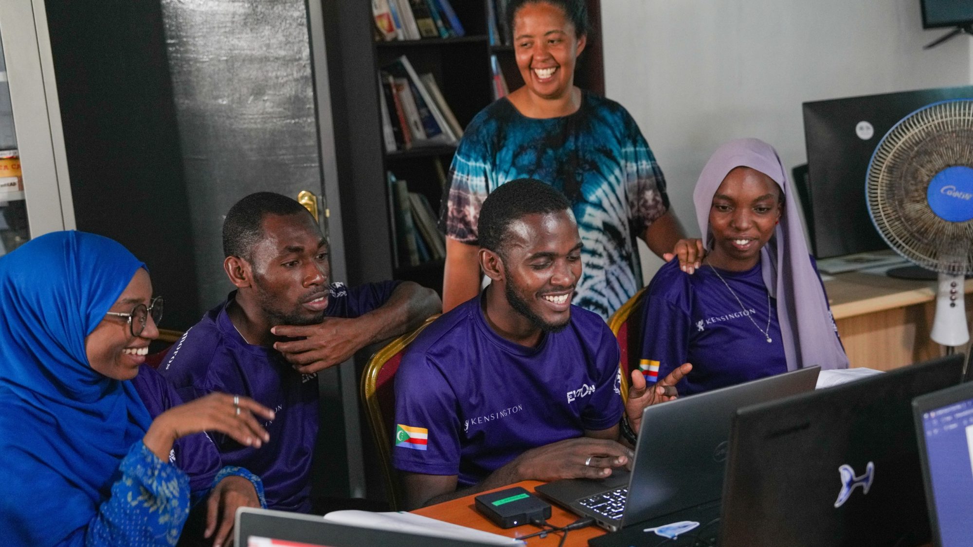 A lively group in Comoros participates in a knowledge exchange session, sharing smiles and ideas. Several people, including a woman wearing a hijab, are gathered around laptops, while a facilitator in the background encourages collaboration in a vibrant learning environment.